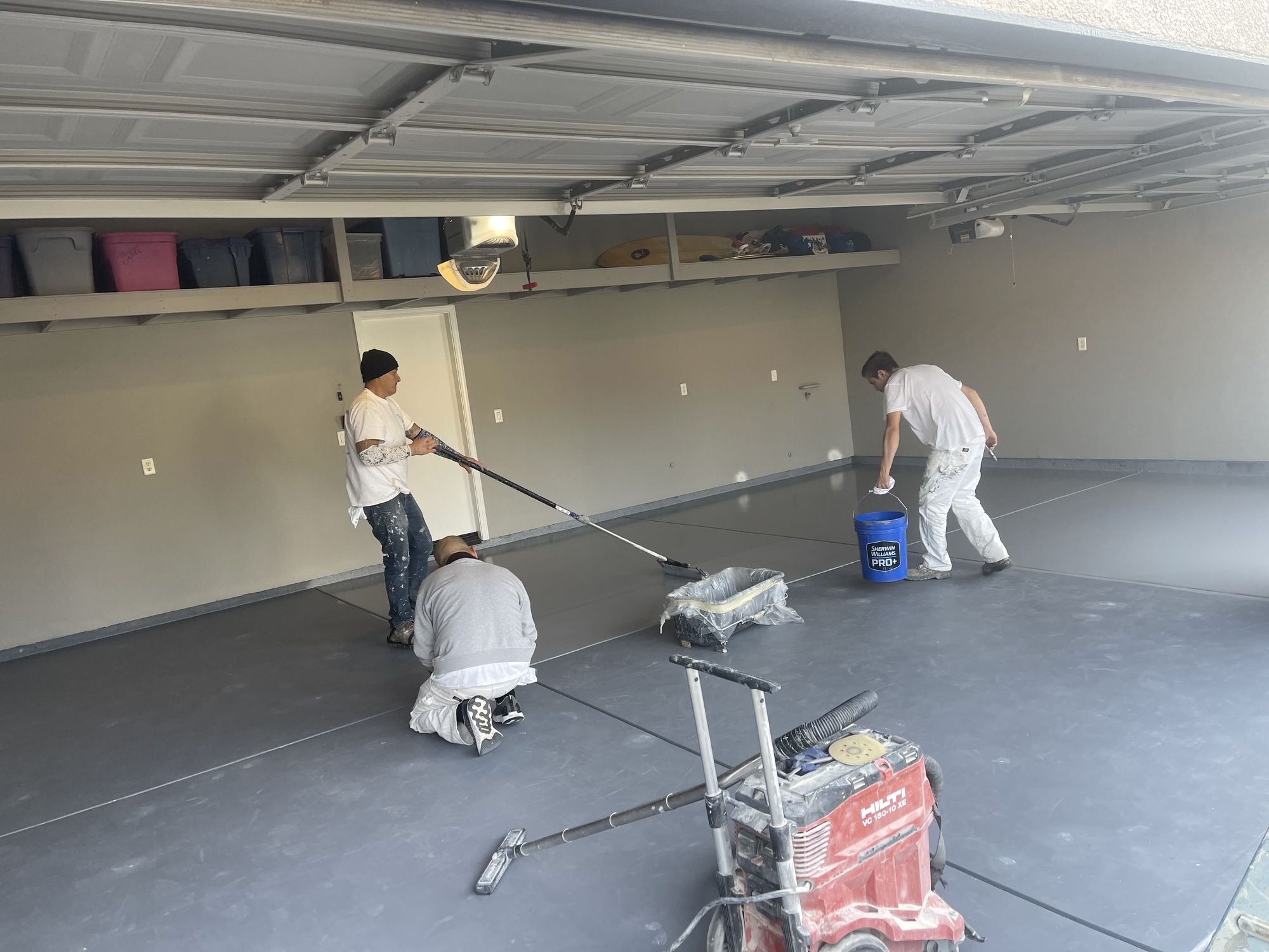 Crew rolling a solid gray garage-floor coating, with a grinder and vacuum on the slab