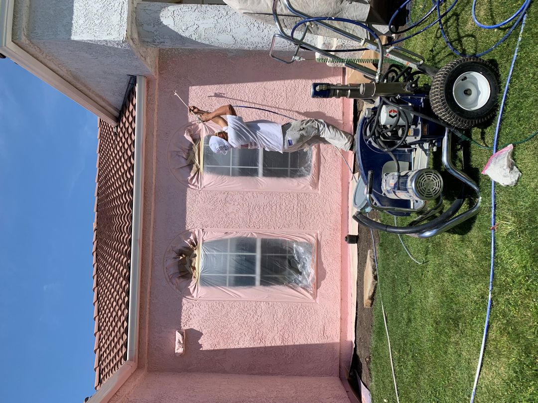 Pink stucco being sprayed, with a crew member on a ladder and masking still on the windows
